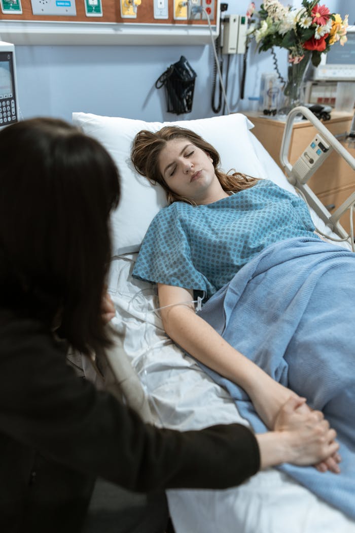 Caucasian woman resting in hospital bed being comforted by visitor holding her hand.